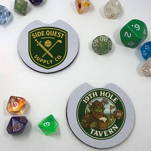 Op de afbeelding: Twee auto-onderzetters met gaming-thema logo's en verschillende polyhedrale dobbelstenen. Eén onderzetter toont de tekst "SIDE QUEST SUPPLY CO." in een schildontwerp. De andere heeft een cartoon van een groenhuidig personage met de tekst "19TH HOLE TAVERN".