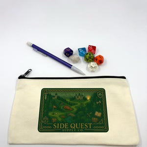 May include: A canvas pencil case with a black zipper, featuring a green and gold graphic of a fantasy landscape with the text "SIDE QUEST SUPPLY CO." Several polyhedral dice and a purple pen are next to the case.