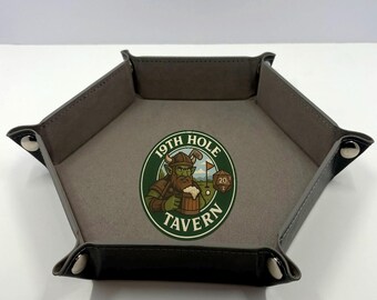 May include: Hexagonal dice tray with a gray interior and black exterior. The tray features a circular graphic with the text "19th Hole Tavern" and an illustration of a green-skinned character holding a mug of beer. The tray is secured with silver rivets.