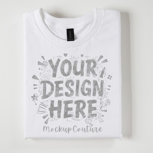 May include: A folded white t-shirt with the text "YOUR DESIGN HERE" in a silver, distressed font. The shirt has a black Gildan tag at the collar. The words "Mockup Couture" are printed below the main text.