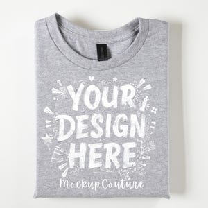 May include: Folded gray t-shirt with the text "YOUR DESIGN HERE" in white, surrounded by decorative elements. The words "Mockup Couture" are at the bottom. The shirt has a crew neck and a black tag.