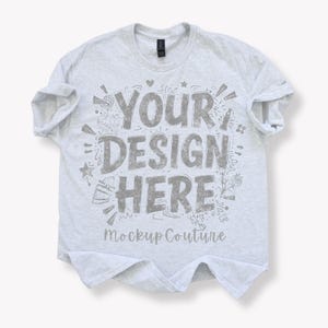 Könnte beinhalten: Ein hellgraues T-Shirt mit hochgekrempelten Ärmeln. Das Shirt zeigt den Text "YOUR DESIGN HERE" in einer stilisierten Schriftart, umgeben von dekorativen Elementen. Die Worte "Mockup Couture" sind unter dem Haupttext aufgedruckt.