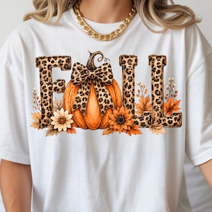 May include: A white t-shirt featuring a graphic print of the word FALL in a leopard print pattern. The letter A is replaced by an orange pumpkin with a leopard print bow. The design includes orange sunflowers and autumn leaves at the base.
