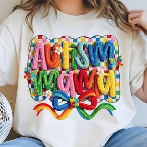 May include: Cream-colored t-shirt with the words "AUTISM MAMA" in colorful, textured letters. The design includes a rainbow bow, daisies, and puzzle piece accents, set against a checkered background.