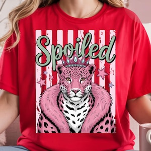 Op de afbeelding: Rood T-shirt met een roze luipaard met een kroon en een roze bontkraag. Het woord "Spoiled" staat in groene letters boven de luipaard. De achtergrond heeft verticale witte en roze strepen en roze sterren.