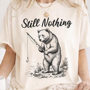 Könnte beinhalten: Cremefarbenes T-Shirt mit dem Text "Still Nothing" über einer Schwarz-Weiß-Illustration eines Bären beim Angeln. Der Bär hält eine Angelrute mit Haken und steht in der Nähe von Wasser und Felsen.