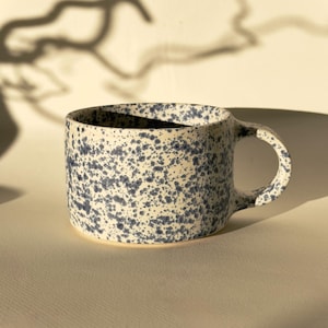 May include: A ceramic mug with a speckled design. The mug is off-white with blue speckles and a rounded handle. The mug is sitting on a light-colored surface.