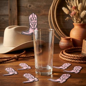 Könnte beinhalten: Ein klares Glas mit rosa Cowboy-Stiefel-förmigen Getränkerührern auf einem Holztisch. Die Rührer tragen Namen. Ein Cowboyhut, ein Seil und Töpferwaren befinden sich im Hintergrund. Die Rührer sind etwa 7.6 cm hoch.