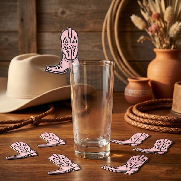 May include: A clear glass with pink cowboy boot-shaped drink stirrers on a wooden table. The stirrers have names on them. A cowboy hat, rope, and pottery are in the background. The stirrers are approximately 3 inches tall.