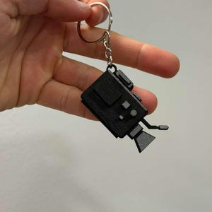 May include: A black keychain featuring a miniature video camera design. The camera has detailed features, including a lens, viewfinder, and buttons. The keychain is attached to a silver metal ring and chain.