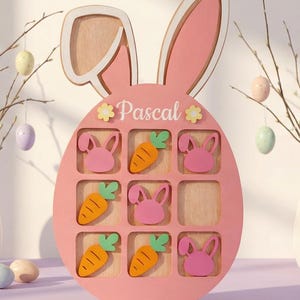 May include: Pink wooden Easter-themed display with bunny ears and an egg shape. It features nine compartments with bunny and carrot cutouts. The name "Pascal" is written on the front. Decorative Easter eggs hang in the background.