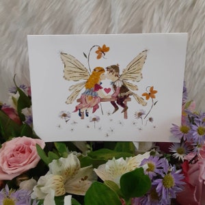 May include: A greeting card with a floral illustration of two fairies kissing on a bench. The fairies have wings made of dried flowers and are surrounded by small white flowers.