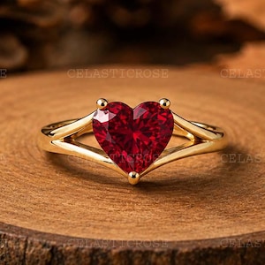 May include: A gold ring featuring a heart-shaped red gemstone set in a split shank band. The ring is positioned on a circular wooden slice. The text CELASTICROSE is repeated across the image in a watermark pattern.