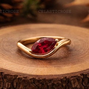 Op de afbeelding: Een gouden ring met een marquise-geslepen, dieprode edelsteen. De ring heeft een vloeiend, asymmetrisch ontwerp. De ring wordt gepresenteerd op een rustiek houten oppervlak, met een vage achtergrond van dennenappels en bladeren.