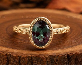 May include: A gold ring featuring an oval-cut, color-changing gemstone that shifts between deep green and purple hues. The band is crafted from yellow gold and displays intricate, embossed floral detailing along the sides of the setting.