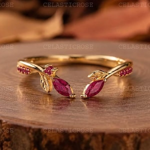 May include: An open-style gold ring featuring two marquise-cut red gemstones at the ends, accented by small red stones along the band. The design incorporates leaf-like metal details surrounding the primary red stones, creating a botanical aesthetic.