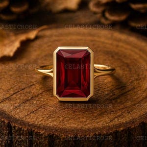 May include: A gold ring with a rectangular, deep red gemstone. The ring is set on a wooden surface, with pine cones and leaves in the background. The ring's design is simple, with a gold band and a bezel setting for the gemstone.
