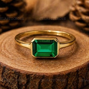 Könnte beinhalten: Ein goldener Ring mit einem rechteckigen, smaragdgeschliffenen grünen Edelstein. Der Ring liegt auf einer Holzoberfläche mit natürlichem, rustikalem Aussehen. Der Hintergrund umfasst Tannenzapfen und Blätter.