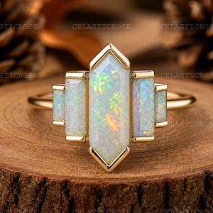 Peut inclure: Une bague en or ornée d'une opale centrale allongée aux couleurs irisées, flanquée de petites opales rectangulaires. La bague présente un design géométrique, posée sur une surface en bois. La bague est un bijou.