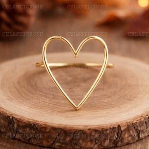 Puede incluir: Un anillo dorado con un diseño en forma de corazón. El anillo está hecho de una fina banda de metal que forma el contorno de un corazón. El anillo se muestra sobre una superficie de madera.