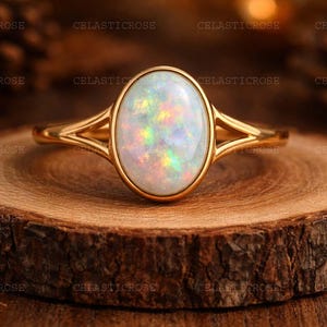 Puede incluir: Un anillo de oro con una piedra preciosa de ópalo ovalada. El ópalo tiene colores iridiscentes de verde, amarillo y rosa. El anillo está sobre una superficie de madera con veta natural.