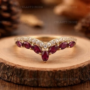 Könnte beinhalten: Ein goldener Ring mit V-förmigem Design, besetzt mit oval geschliffenen Rubin-Edelsteinen und kleinen, runden Diamanten. Der Ring wird auf einer Holzscheibe präsentiert, die die Handwerkskunst und Eleganz des Schmucks hervorhebt.