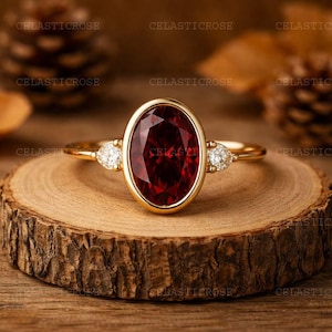 Könnte beinhalten: Ein goldener Ring mit einem großen, oval geschliffenen, tiefroten Edelstein. Zwei kleinere, birnenförmige, klare Edelsteine flankieren den zentralen Stein. Der Ring liegt auf einer Holzoberfläche, die das Design und die Farbe des Schmucks hervorhebt.