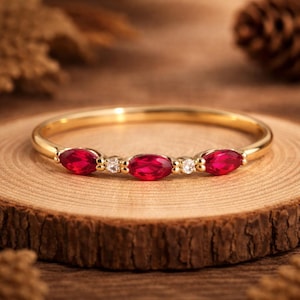 Puede incluir: Un anillo de oro con cinco piedras preciosas rojas ovaladas, intercaladas con pequeñas piedras blancas. El anillo se exhibe sobre una superficie de madera, resaltando su diseño elegante. La banda del anillo es de oro pulido.