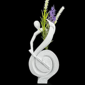 Peut inclure: Un vase sculpture abstrait blanc moucheté en forme de clé de sol avec une silhouette humaine stylisée intégrée au design. Le vase contient un petit arrangement de tiges florales artificielles vertes, blanches et violettes.