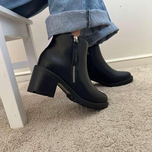 May include: Black ankle boots with a chunky heel and a side zipper. The boots have a rounded toe and a lug sole. The person is wearing blue jeans. The boots are sitting on a beige carpet.
