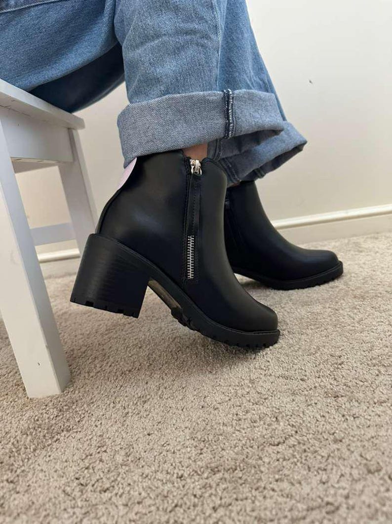 May include: Black ankle boots with a chunky heel and a side zipper. The boots have a rounded toe and a lug sole. The person is wearing blue jeans. The boots are sitting on a beige carpet.