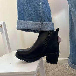 May include: Black ankle boot with a chunky heel and a side zipper. The boot has a rounded toe and a lug sole. The boot is worn with blue jeans.