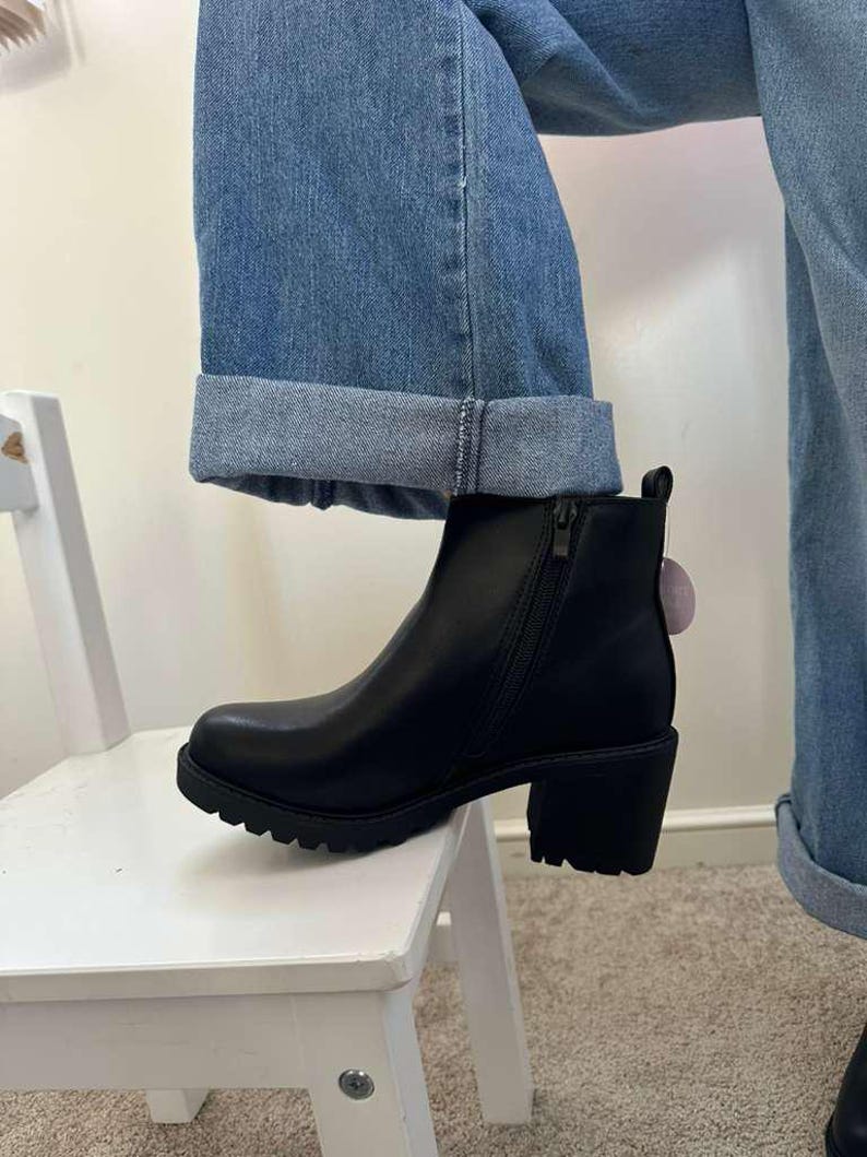 May include: Black ankle boot with a chunky heel and a side zipper. The boot has a rounded toe and a lug sole. The boot is worn with blue jeans.