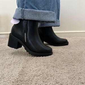 May include: Black ankle boots with a chunky heel and a side zipper. The boots have a round toe and a lug sole. The boots are worn with blue jeans.