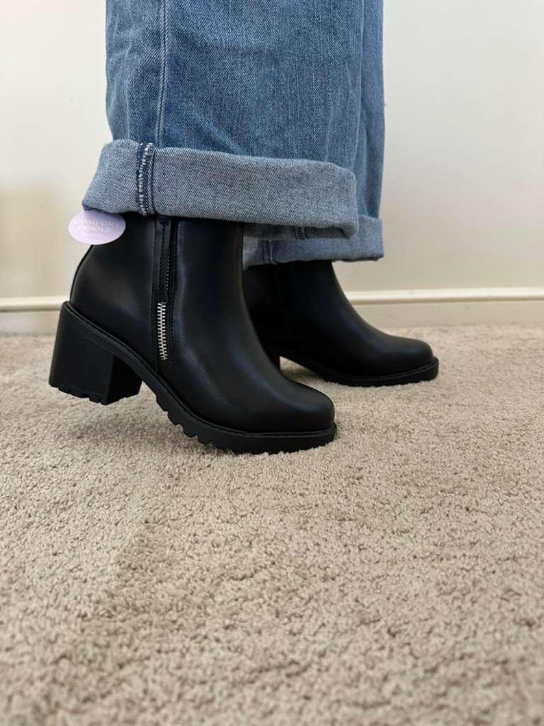 May include: Black ankle boots with a chunky heel and a side zipper. The boots have a round toe and a lug sole. The boots are worn with blue jeans.