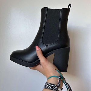 May include: A black ankle boot with a block heel is held up by a hand. The boot has a rounded toe and elastic side panels. The heel is approximately 3 inches high. The boot is made of a smooth, matte material.