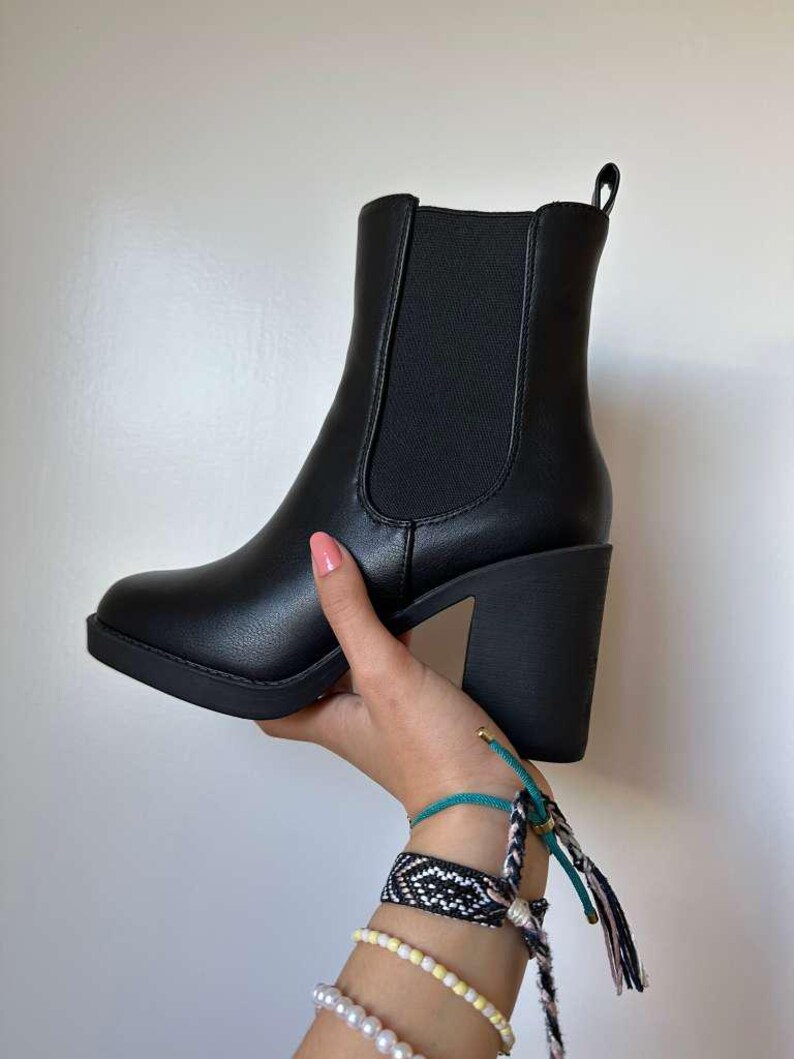 May include: A black ankle boot with a block heel is held up by a hand. The boot has a rounded toe and elastic side panels. The heel is approximately 3 inches high. The boot is made of a smooth, matte material.