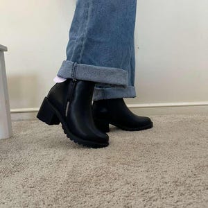 May include: Black ankle boots with a block heel and a side zipper. The boots have a rounded toe and a textured sole. The boots are worn with blue denim jeans with a rolled cuff.