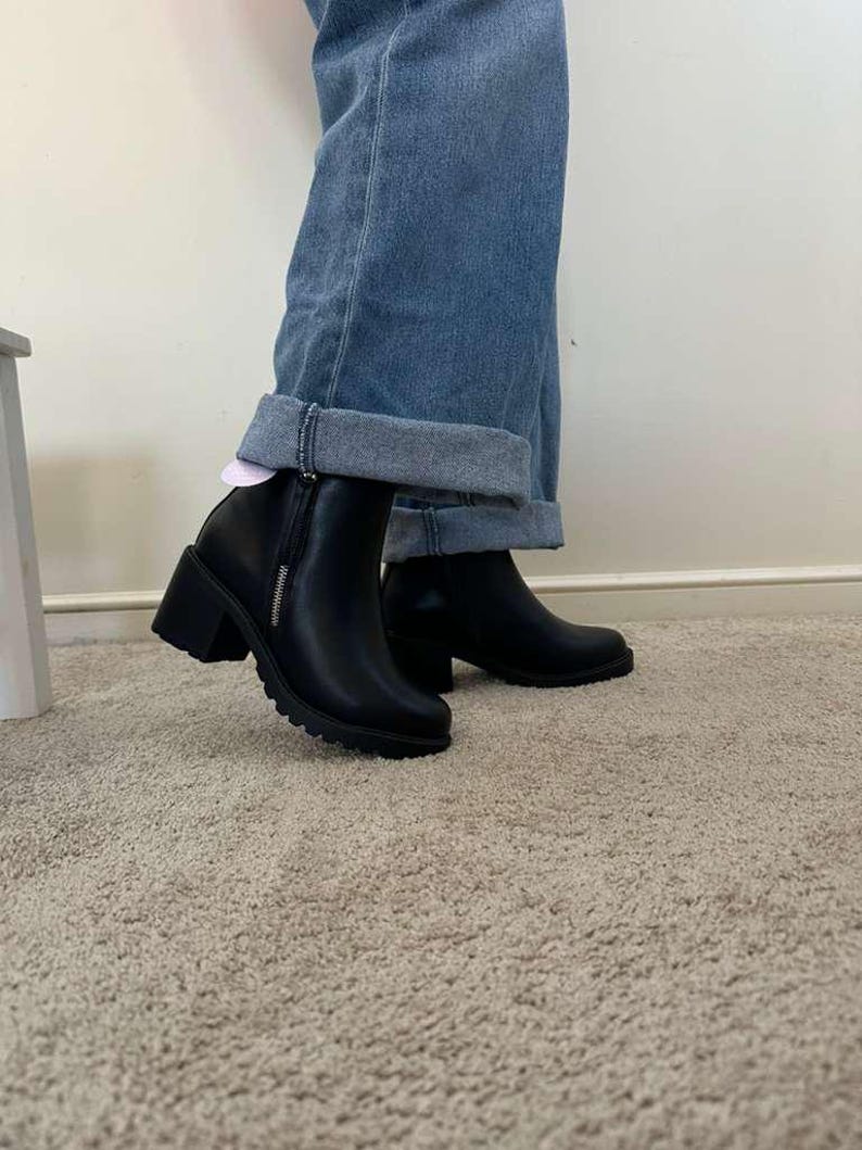 May include: Black ankle boots with a block heel and a side zipper. The boots have a rounded toe and a textured sole. The boots are worn with blue denim jeans with a rolled cuff.
