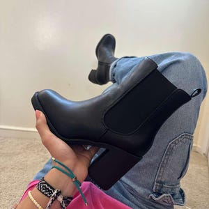 May include: Black ankle boots with a block heel. The boots have a smooth leather-like appearance and elastic side panels. The heel is approximately 3 inches high. The boots are shown with blue jeans and a pink top.