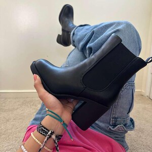 May include: Black ankle boots with a block heel. The boots have a rounded toe and elastic side panels. The person is wearing blue jeans and a pink shirt. The boots are a classic style.