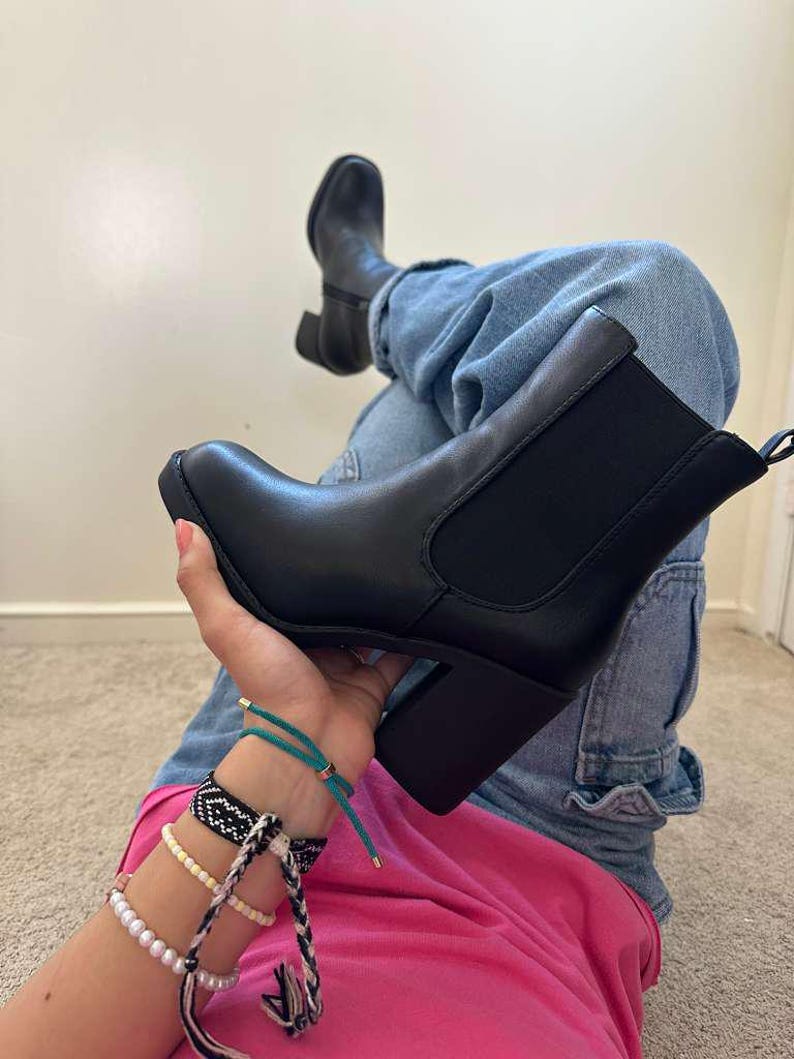 May include: Black ankle boots with a block heel. The boots have a rounded toe and elastic side panels. The person is wearing blue jeans and a pink shirt. The boots are a classic style.