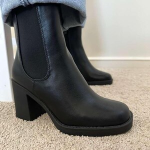 May include: Black ankle boots with a block heel and rounded toe. The boots have a Chelsea boot design with elastic side panels. The boots are made of a smooth material and are worn with blue jeans.