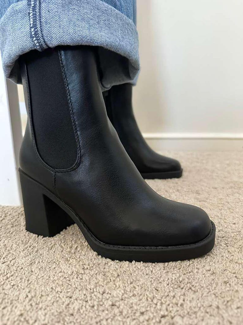 May include: Black ankle boots with a block heel and rounded toe. The boots have a Chelsea boot design with elastic side panels. The boots are made of a smooth material and are worn with blue jeans.