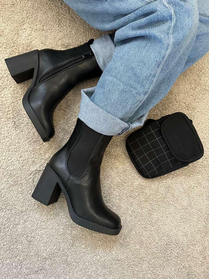 May include: Black leather ankle boots with block heels and a side zipper. The boots have a rounded toe and a platform sole. A small black square wallet with a grid pattern is next to the boots.