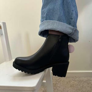 May include: Black ankle boot with a chunky heel and a lug sole. The boot has a side zipper and a small tag attached. The boot is worn with blue jeans.