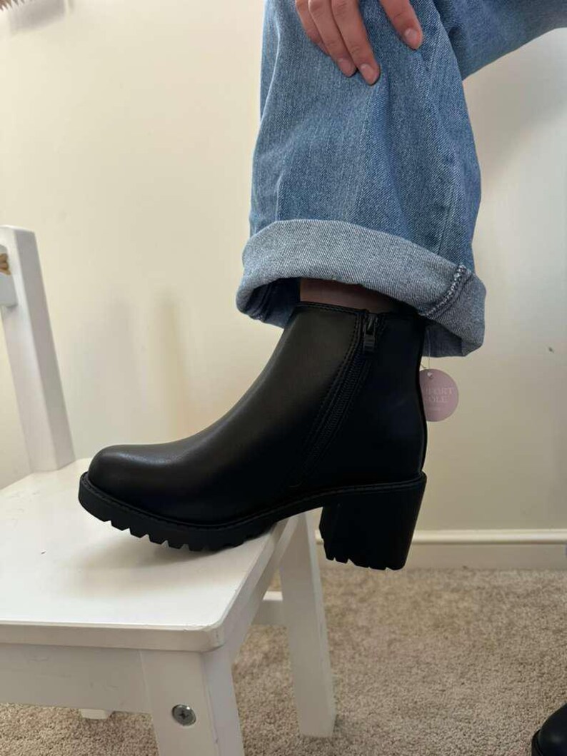 May include: Black ankle boot with a chunky heel and a lug sole. The boot has a side zipper and a small tag attached. The boot is worn with blue jeans.