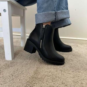 May include: Black ankle boots with a chunky heel and a zipper closure. The boots have a rounded toe and a textured sole. The boots are worn with blue jeans.
