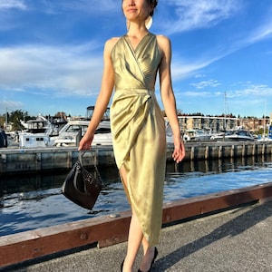 May include: A gold-colored, halter-style dress with a wrap design and a high slit. The dress is paired with black heels and a black handbag with a chain strap. The background features a marina with boats and a blue sky.