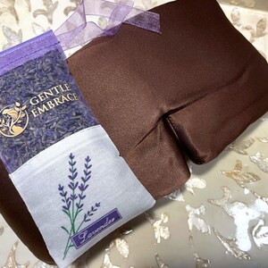 May include: A brown eye pillow and a lavender sachet. The eye pillow is rectangular and made of a smooth fabric. The sachet is filled with lavender buds and has the text "GENTLE EMBRACE" and a lavender sprig design. A purple ribbon is attached.